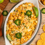 A baked street corn dip in a white oval dish, topped with melted cheese, sliced jalape&ntilde;os, chopped cilantro, and lime wedges, surrounded by tortilla chips and fresh limes.