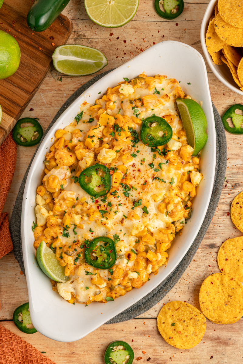 A baked street corn dip in a white oval dish, topped with melted cheese, sliced jalapeños, chopped cilantro, and lime wedges, surrounded by tortilla chips and fresh limes.