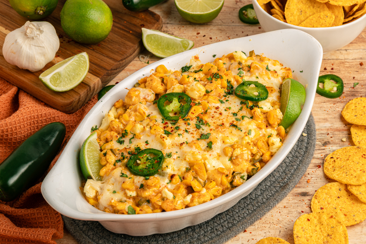 Street corn dip in a white casserole dish, garnished with jalapeños, cilantro, and lime wedges, with fresh limes, jalapeños, and tortilla chips arranged around it.