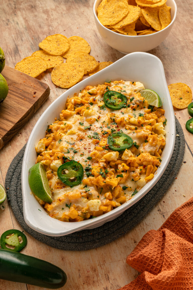 An overhead view of cheesy Mexican corn dip served in a white baking dish, surrounded by tortilla chips and fresh lime wedges.
