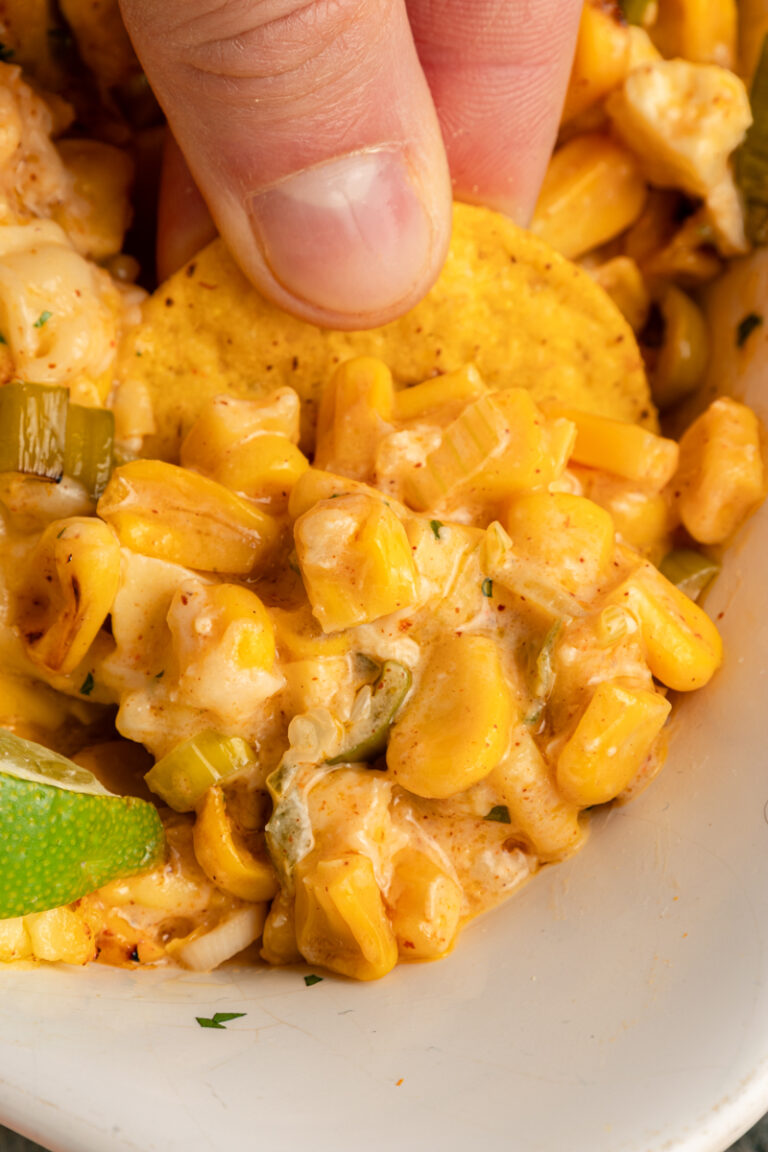 A close-up of tortilla chips being dipped into warm street corn dip with corn kernels, cheese, jalapeño pieces, and lime visible in the dish.