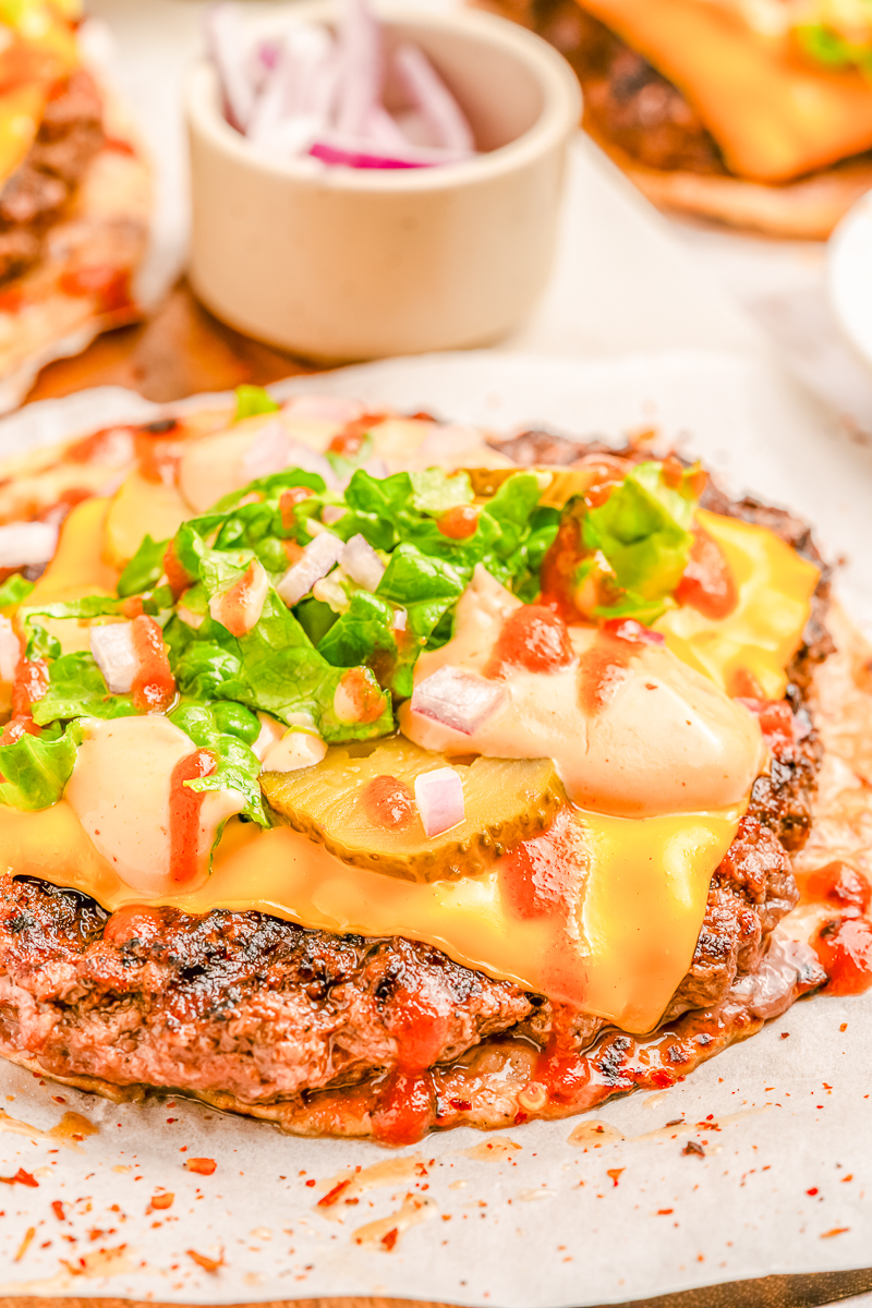 Angle shot of a beef-topped tortilla layered with cheddar cheese, pickles, lettuce, onions, and creamy sauce, with the sauce bowl in the background.
