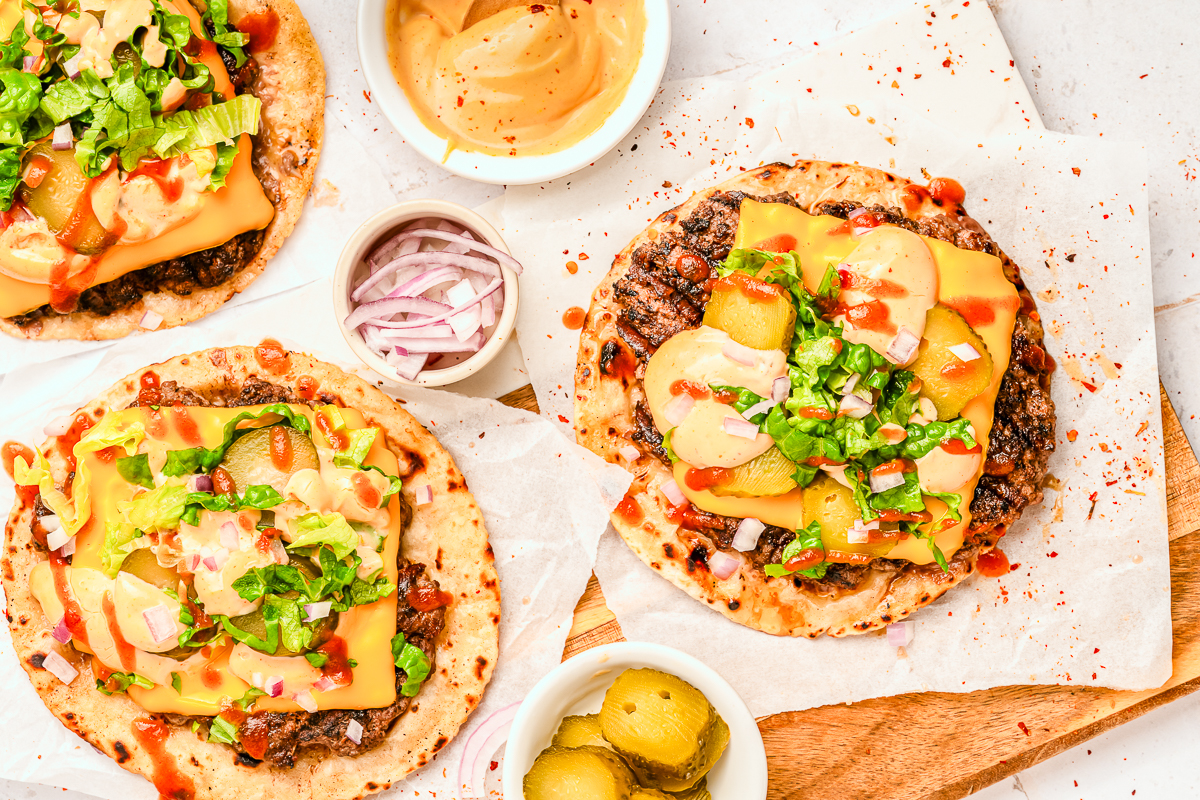 Overhead arrangement of tortillas topped with beef, cheddar, pickles, lettuce, onions, and sauce, with small bowls of extra toppings and a sauce bowl for the smash burger tacos.