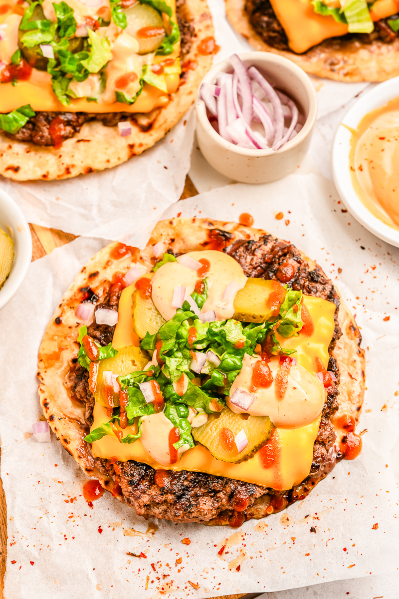 Overhead view of a tortilla loaded with seared beef, melted cheddar, pickles, lettuce, red onion, and creamy sauce, with a bowl of dipping sauce nearby.